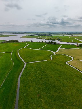 Aerial view of a wide green landscape with winding canals dividing the fields. Cows graze on the pastures, and a large river flows in the distance under a cloudy sky. Peaceful rural scenery.