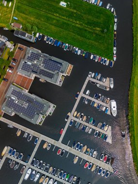 Ahşap rıhtımlar boyunca kenetlenmiş birden fazla teknesi olan bir marinanın tepeden aşağı manzarası, yeşil parklar ve doğal çimenlik alanlarla çevrili dar bir su yolunda seyreden bir tekne. Güneş panelleri.