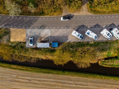 Açık mavi bir gökyüzünün altında, ufka doğru uzanan manzaralı ormanlık bir arazide, yol kenarına park edilmiş karavan manzarası..