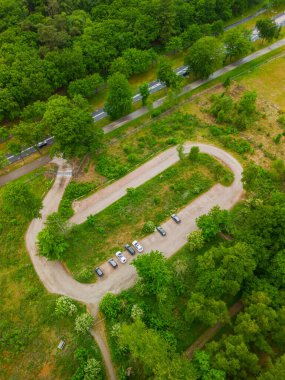 Çevresi yemyeşil ağaçlar ve çayırlarla çevrili park halindeki küçük, kıvrımlı bir orman otoparkının havadan görüntüsü, görüntünün üst kısmından geçen bir yol..