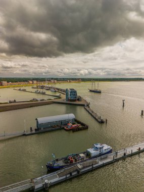 Dramatik bulutlu gökyüzünün altında geniş bir su kütlesi üzerinde birkaç tekne ve geminin bulunduğu bir liman manzarası, modern bir rıhtım binası ve ön planda görünen rıhtımlar var..