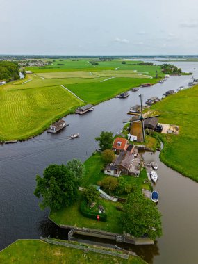 Çiçekli yeşil tarlalar, geleneksel evler ve bulutlu bir gökyüzünün altındaki uzak su yollarıyla çevrili bir kanalın yanındaki Hollandalı yel değirmeni manzarası..