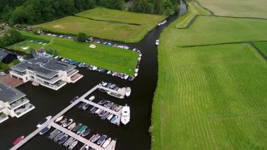 Ahşap rıhtımlar boyunca kenetlenmiş bir marina manzarası, yeşil parklar ve doğal otlaklarla çevrili dar bir su yolunda seyreden bir tekne..