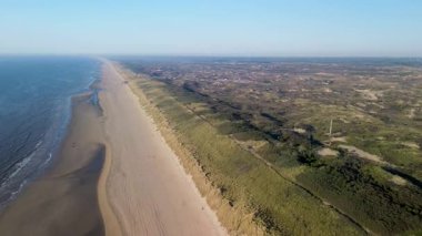 Sahil kulübeleriyle dolu güzel bir kumsal manzarası, kıyıda dinlenen insanlar ve ön planda çimenli kum tepeleriyle çevrili, kıyıya vuran nazik dalgalar..