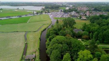 Dar bir su kanalı çevreleyen ağaçları yansıtan yeşil bir ormandan geçiyor. Arka planda güzel bir Hollanda köyü var. Yatı olan büyük bir iskele. Yeşil bir çayır, bir tarla.