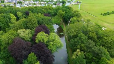 Bereketli ağaçlarla dolu huzurlu bir parkın havadan görünüşü sakin su üzerinde bir yaya köprüsü ve yürüyüş yolları. Yakınlarda, beyaz evleri ve ötesine uzanan yeşil tarlaları olan bir kasabaya giden bir kanal var..