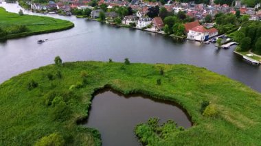 Bereketli sulak arazileri ve kalp şeklinde göleti olan bir nehir manzarası. Rıhtım boyunca yanaşmış tekneleri ve yanından geçen huzurlu bir teknesi olan büyüleyici bir yerleşim yerine bakıyor..