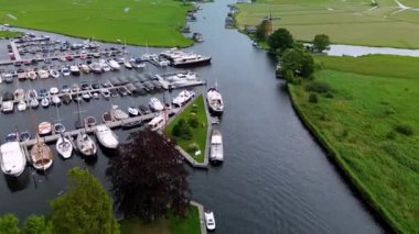 Ahşap rıhtımlar boyunca kenetlenmiş bir marina manzarası, yeşil parklar ve doğal otlaklarla çevrili dar bir su yolunda seyreden bir tekne..