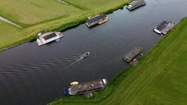 Uçsuz bucaksız yeşil çayırlar ve kesişen su yollarıyla çevrili nehirde yüzen evlerin sakin bir görüntüsü. Hollanda bayrakları bulutlu bir gökyüzünün altında rüzgarda hafifçe sallanıyor..