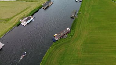 Uçsuz bucaksız yeşil çayırlar ve kesişen su yollarıyla çevrili nehirde yüzen evlerin sakin bir görüntüsü. Hollanda bayrakları bulutlu bir gökyüzünün altında rüzgarda hafifçe sallanıyor..