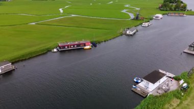 Uçsuz bucaksız yeşil çayırlar ve kesişen su yollarıyla çevrili nehirde yüzen evlerin sakin bir görüntüsü. Hollanda bayrakları bulutlu bir gökyüzünün altında rüzgarda hafifçe sallanıyor..