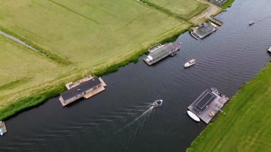 Uçsuz bucaksız yeşil çayırlar ve kesişen su yollarıyla çevrili nehirde yüzen evlerin sakin bir görüntüsü. Hollanda bayrakları bulutlu bir gökyüzünün altında rüzgarda hafifçe sallanıyor..