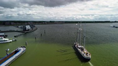 Dramatik bulutlu gökyüzünün altında geniş bir su kütlesi üzerinde birkaç tekne ve geminin bulunduğu bir liman manzarası, modern bir rıhtım binası ve ön planda görünen rıhtımlar var..