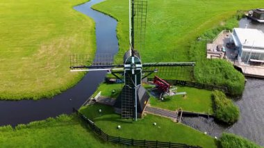 Hollanda 'nın klasik yel değirmeni, dolambaçlı bir kanalın yanındaki yemyeşil tarlaların ortasında duruyor. Geleneksel yapı huzurlu kırsal kesimle çevrili, kırsal çekicilik sergiliyor..