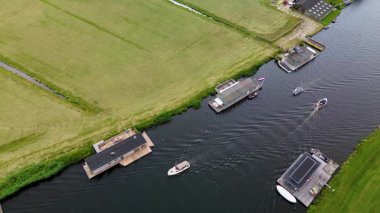 Uçsuz bucaksız yeşil çayırlar ve kesişen su yollarıyla çevrili nehirde yüzen evlerin sakin bir görüntüsü. Hollanda bayrakları bulutlu bir gökyüzünün altında rüzgarda hafifçe sallanıyor..