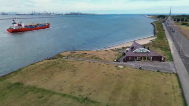 Endüstriyel bir liman yakınındaki geniş bir nehir veya kanalda ilerleyen kargo gemilerinin hava görüntüsü açık mavi gökyüzünün altında vinçler ve bacalar var..