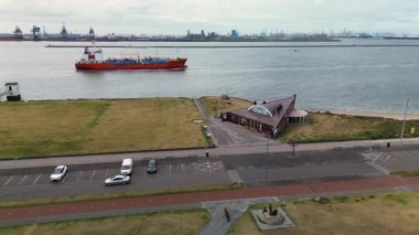 Endüstriyel bir liman yakınındaki geniş bir nehir veya kanalda ilerleyen kargo gemilerinin hava görüntüsü açık mavi gökyüzünün altında vinçler ve bacalar var..