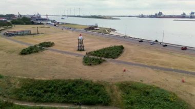 Endüstriyel bir liman yakınındaki geniş bir nehir ya da kanalda seyreden kargo gemilerinin arka planda bulutlu bir gökyüzünün altında vinçleri ve duman yığınları var..