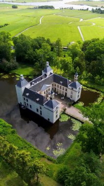 Uçsuz bucaksız yeşil tarlaları, dolambaçlı su yolları ve Hollanda 'nın düz kırsalında parçalı bulutlu bir gökyüzünün altında uzanan bir nehirle çevrili tarihi bir şatonun havadan görünüşü..