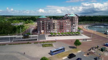 Büyük tuğladan bir apartman dairesi, rıhtımda gezinti alanı, ağaçlarla kaplı caddeler ve parçalı bulutlu bir gökyüzünün altında bir marina ile manzaralı bir drone görüntüsü..