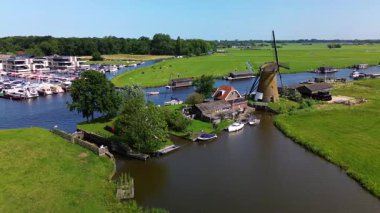 Warmond, Güney Hollanda, Hollanda - 09.06.2025: Kanal boyunca beyaz yelkenli bir yat. Güzel bir yel değirmeni. Dar kanalın havadan görünüşü yemyeşil çayırlarda ve küçük bir ormanda akıyor.