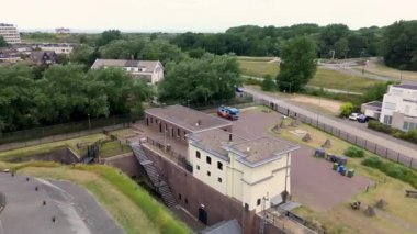 Modern binalar, ağaçlar ve yürüyüş yolları ile çevrili çimenlerle kaplı tarihi bir askeri kalenin havadan görünüşü, kıyı şeridi yakınlarında parlak mavi bir gökyüzünün altında..