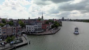 Kubbeli bir kulesi, geleneksel binaları, ağaçlarla kaplı caddeleri ve bulutlu bir gökyüzünün altında geniş bir nehirde yelken açan bir teknesi olan tarihi bir Avrupa şehri manzarası..