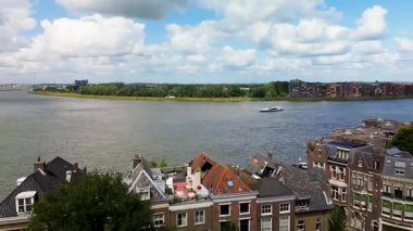 Dik çatıları olan geleneksel Hollanda evlerinin manzaralı nehir manzarası, sudan geçen bir feribot, ve arkaplanda parçalı bulutlu bir gökyüzünün altında modern binalar ve yeşillikler..