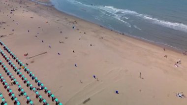 Sahil kulübeleriyle dolu güzel bir kumsal manzarası, kıyıda dinlenen insanlar ve ön planda çimenli kum tepeleriyle çevrili, kıyıya vuran nazik dalgalar..