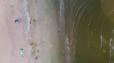 Sahil kulübeleriyle dolu güzel bir kumsal manzarası, kıyıda dinlenen insanlar ve ön planda çimenli kum tepeleriyle çevrili, kıyıya vuran nazik dalgalar..