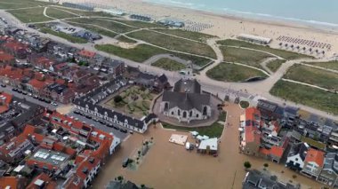 Kırmızı çatılı evleri olan bir sahil kasabasının havadan görünüşü, tarihi bir kilise, kum tepeleri boyunca kıvrılan yollar, ve kıyı şeridi boyunca şemsiyeler ve plaj kulübeleriyle kaplı geniş kumlu bir sahil..