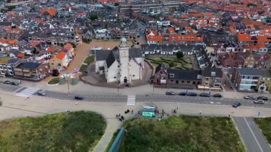 Kırmızı çatılı evleri, merkezi bir kilisesi ve geometrik bahçesi olan Avrupa kıyı kasabasının havadan görünüşü. Arabalı geniş bir yol ve kum tepeciklerinin yanında ön planda bisiklet şeridi var..