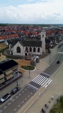 Kırmızı çatılı evleri, merkezi bir kilisesi ve geometrik bahçesi olan Avrupa kıyı kasabasının havadan görünüşü. Arabalı geniş bir yol ve kum tepeciklerinin yanında ön planda bisiklet şeridi var..