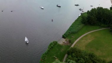 Bereketli yeşilliklerle çevrili sularda yüzen yelkenli ve yatlarla dolu huzurlu bir nehir manzarası ve ağaçlar ve çimenler arasında dolambaçlı bir yol..