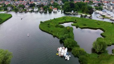 Ahşap rıhtımlar boyunca kenetlenmiş bir marina manzarası, yeşil parklar ve doğal otlaklarla çevrili dar bir su yolunda seyreden bir tekne..