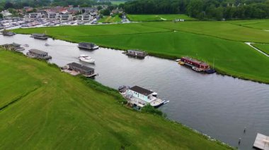 Uçsuz bucaksız yeşil çayırlar ve kesişen su yollarıyla çevrili nehirde yüzen evlerin sakin bir görüntüsü. Hollanda bayrakları bulutlu bir gökyüzünün altında rüzgarda hafifçe sallanıyor..