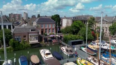 Dik çatıları olan geleneksel Hollanda evlerinin manzaralı nehir manzarası, sudan geçen bir feribot, ve arkaplanda parçalı bulutlu bir gökyüzünün altında modern binalar ve yeşillikler..