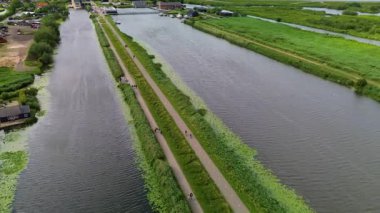 Yürüme yolları olan bir kanal kasabası, kıyı boyunca kenetlenmiş tekneler, yeşil alanlar ve bulutlu gökyüzü. Doğanın ve küçük kasaba hayatının karışımıyla huzurlu bir nehir kenarı manzarası..
