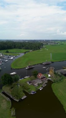Ahşap rıhtımlar boyunca kenetlenmiş birden fazla teknesi olan bir marinanın tepeden aşağı manzarası, yeşil parklar ve doğal çimenlik alanlarla çevrili dar bir su yolunda seyreden bir tekne. Güzel bir rüzgar değirmeni.