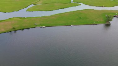 Dar bir kanalın havadan görünüşü yemyeşil çayırların arasından akıyor ve küçük bir orman huzurlu bir Hollanda köyüne götürüyor açık mavi gökyüzünün altında bir marinası ve demirlemiş yatları var..