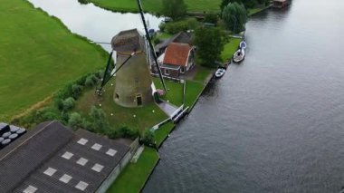 Uçsuz bucaksız yeşil çayırlar ve Hollanda bayraklarıyla kesişen, bulutlu bir gökyüzünün altında rüzgarda hafifçe sallanan su yollarıyla çevrili, nehirde yüzen evlerin sakin bir görüntüsü. Klasik bir Hollanda yel değirmeni