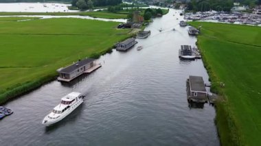Uçsuz bucaksız yeşil çayırlar ve Hollanda bayraklarıyla kesişen, bulutlu bir gökyüzünün altında rüzgarda hafifçe sallanan su yollarıyla çevrili, nehirde yüzen evlerin sakin bir görüntüsü. Klasik bir Hollanda yel değirmeni