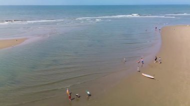 Kumlu bir sahilin havadan görünüşü. İnsanlar sığ gel-git havuzları ve kum havuzlarının yanında yürüyor. Denizden hafif dalgalar yaklaşıyor..