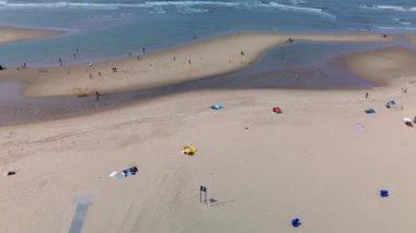 Kumlu bir sahilin havadan görünüşü. İnsanlar sığ gel-git havuzları ve kum havuzlarının yanında yürüyor. Denizden hafif dalgalar yaklaşıyor..