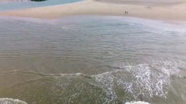 Kumlu bir sahilin havadan görünüşü. İnsanlar sığ gel-git havuzları ve kum havuzlarının yanında yürüyor. Denizden hafif dalgalar yaklaşıyor..