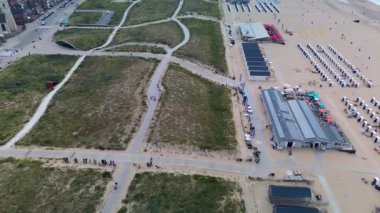 Kumlu bir sahilin havadan görünüşü. İnsanlar sığ gel-git havuzları ve kum havuzlarının yanında yürüyor. Denizden hafif dalgalar yaklaşıyor..