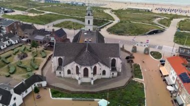 Kumlu bir sahilin havadan görünüşü. İnsanlar sığ gel-git havuzları ve kum havuzlarının yanında yürüyor. Denizden hafif dalgalar yaklaşıyor..