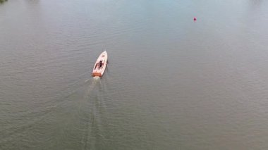 İçinde bir kişi olan küçük beyaz bir motorlu tekne sakin sularda ilerler ve arkalarında hafif bir iz bırakır. Uçsuz bucaksız su, teknenin etrafını sarar. Uzakta tek bir kırmızı şamandıra yüzer..