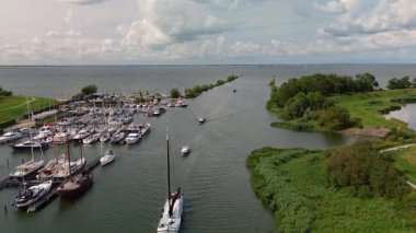 Sayısız yelkenli ve yatla dolu bir marinanın havadan görünüşü geniş bir göle veya denize açılan bir kanal ve parçalı bulutlu bir gökyüzünün altındaki yemyeşil arazi..