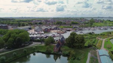 Limanı teknelerle çevrili bir liman kasabasının havadan görüntüsü. Etrafı yeşilliklerle, tarihi binalarla ve parçalı bulutlu bir gökyüzünün altındaki yerleşim yerleriyle çevrili..